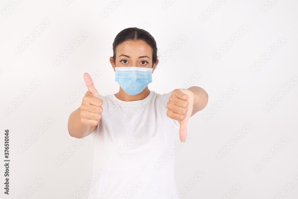 Young arab woman wearing medical mask standing over isolated white background showing thumbs up and thumbs down, difficult choose concept Young arab woman wearing medical mask standing over isolated white background showing thumbs up and thumbs down, difficult choose concept