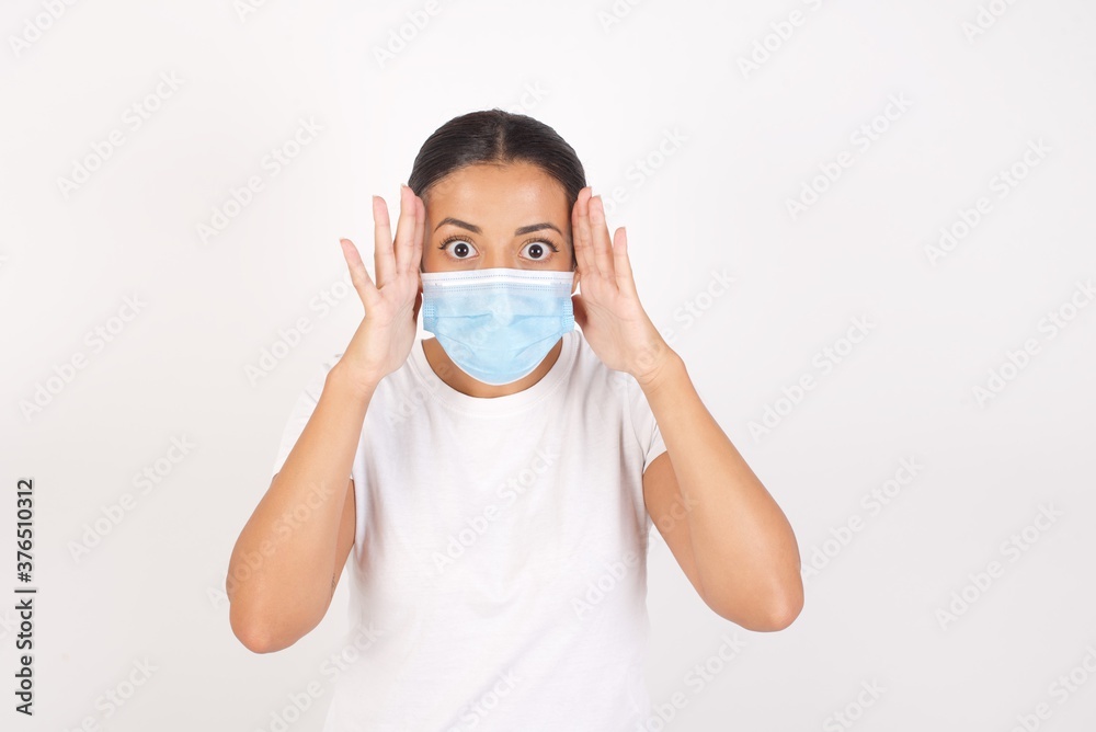 Young arab woman wearing medical mask standing over isolated white background with scared expression, keeps hands on head, jaw dropped, has terrific expression. Omg concept