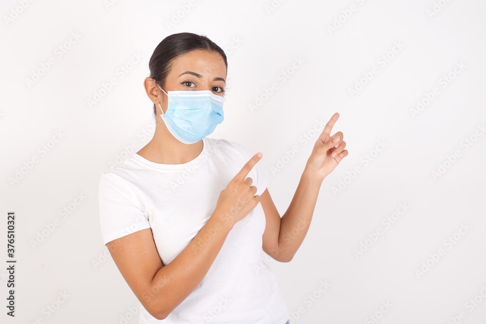 Young arab woman wearing medical mask standing over isolated white background indicating with forefinger empty space showing best low prices Young arab woman wearing medical mask standing over isolated white background indicating with forefinger empty space showing best low prices