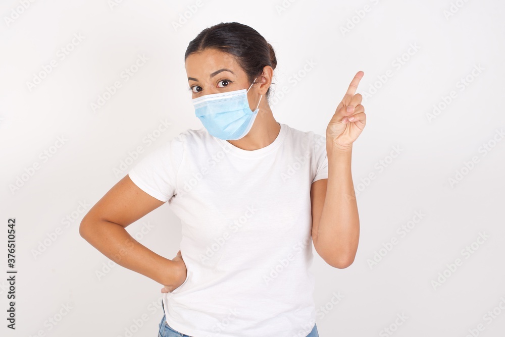Young arab woman wearing medical mask standing over isolated white background holding finger up having idea and posing