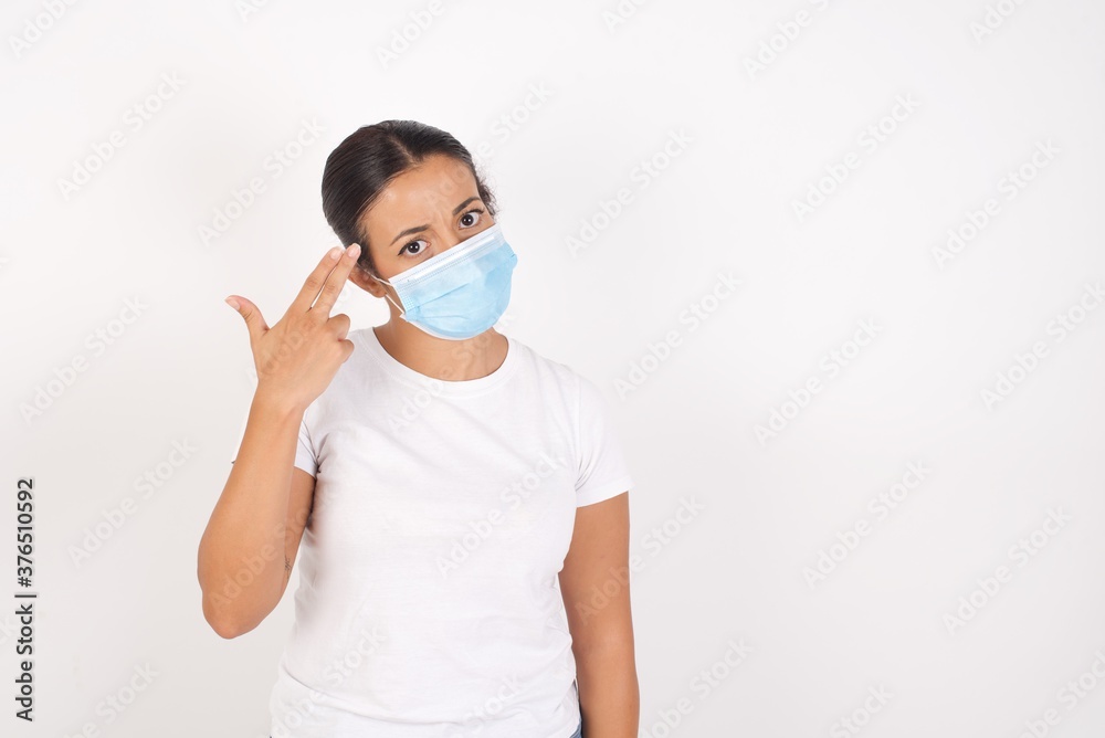 Unhappy Young arab woman wearing medical mask standing over isolated white background makes suicide gesture and imitates gun with hand, curves lips, keeps two fingers on temple