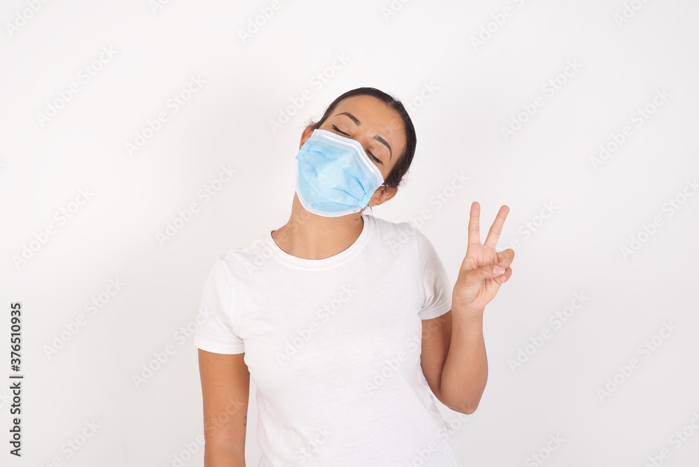 Young arab woman wearing medical mask standing over isolated white background smiling with happy face winking at the camera doing victory sign. Number two. Young arab woman wearing medical mask standing over isolated white background smiling with happy face winking at the camera doing victory sign. Number two.