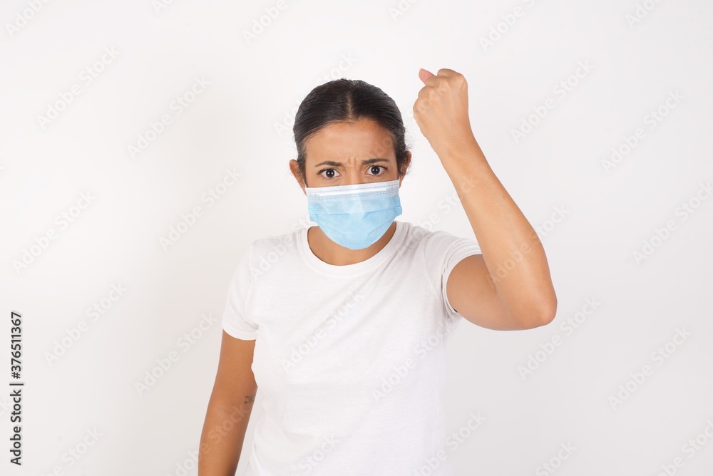 Young arab woman wearing medical mask standing over isolated white background being angry and mad raising fist frustrated and furious while shouting with anger. Rage and aggressive concept. Young arab woman wearing medical mask standing over isolated white background being angry and mad raising fist frustrated and furious while shouting with anger. Rage and aggressive concept.