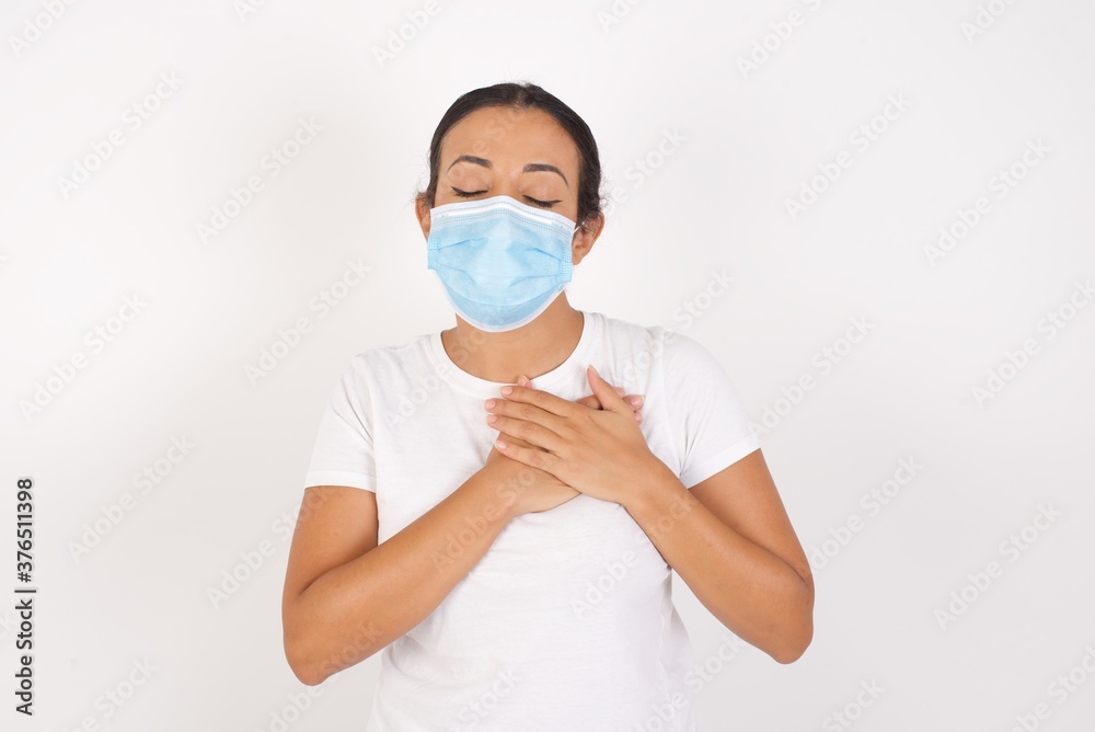 Young arab woman wearing medical mask standing over isolated white background smiling with hands on chest with closed eyes and grateful gesture on face. Health concept.
