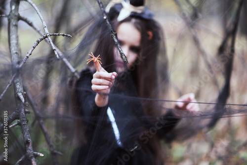 Beautiful little girl, dressed in a witch dress and hattouching spider with her finger in fall forest with fear expressions. Halloween horror. Witches in darkness