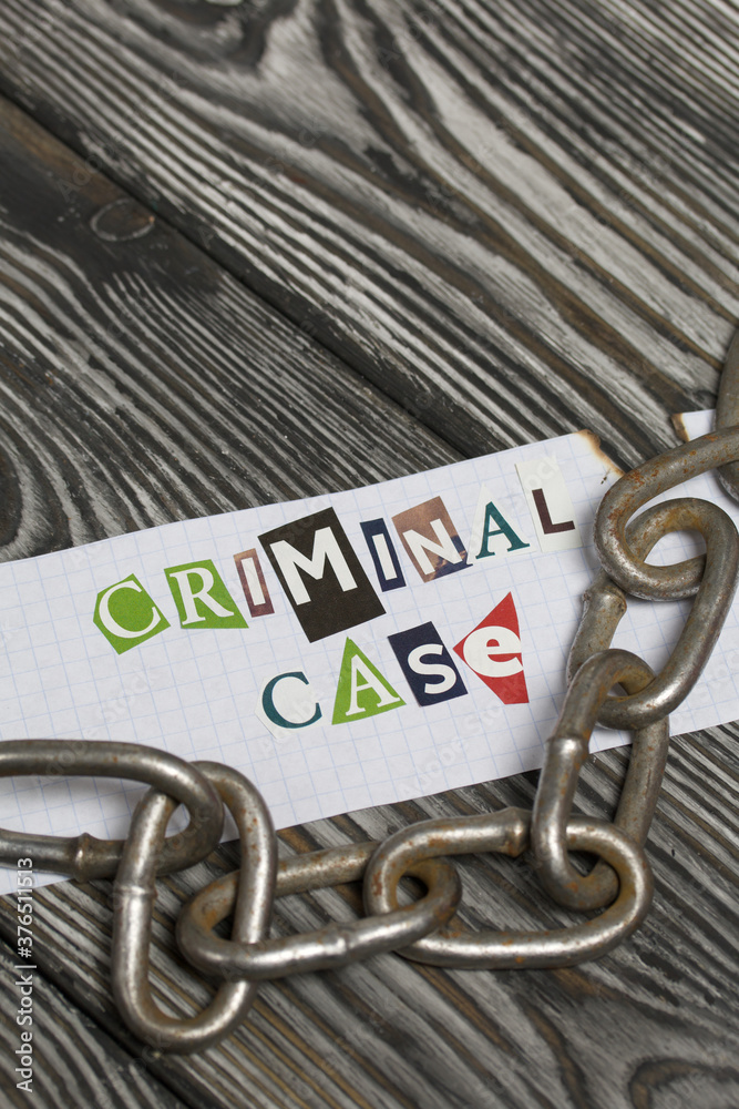 Fototapeta premium The phrase Criminal case made of letters cut from a magazine and pasted on a sheet of paper. There is a metal chain nearby. On brushed pine boards painted black and white.