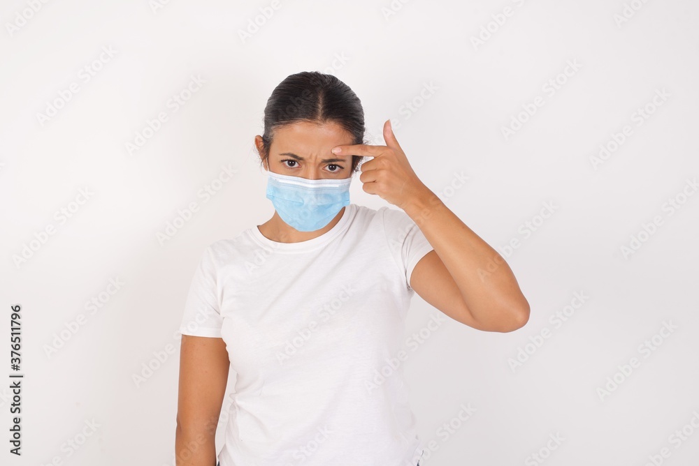 Young arab woman wearing medical mask standing over isolated white background pointing unhappy at pimple on forehead, blackhead infection. Skincare concept. Young arab woman wearing medical mask standing over isolated white background pointing unhappy at pimple on forehead, blackhead infection. Skincare concept.