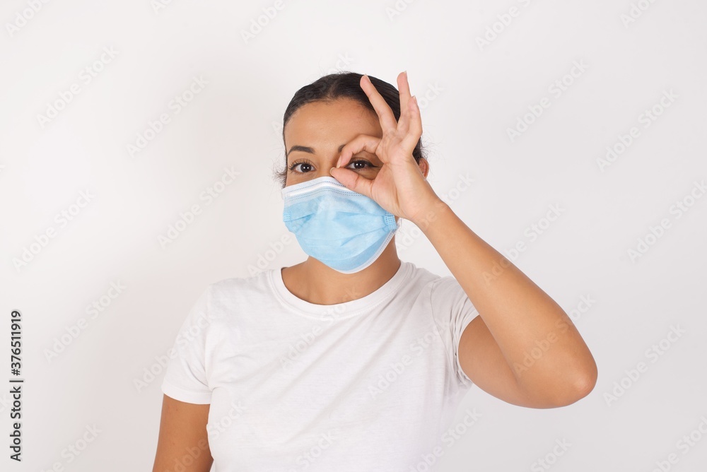 Young arab woman wearing medical mask standing over isolated white background doing ok gesture with hand smiling, eye looking through fingers with happy face. Young arab woman wearing medical mask standing over isolated white background doing ok gesture with hand smiling, eye looking through fingers with happy face.