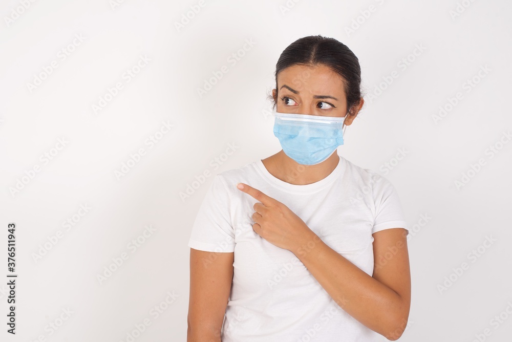 Young arab woman wearing medical mask standing over isolated white background. Pointing aside worried and nervous with forefinger, concern and surprise concept. Young arab woman wearing medical mask standing over isolated white background. Pointing aside worried and nervous with forefinger, concern and surprise concept.
