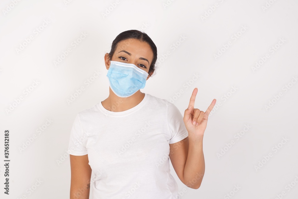 Young arab woman wearing medical mask standing over isolated white background doing a rock gesture and smiling to the camera. Ready to go to her favorite band concert. Young arab woman wearing medical mask standing over isolated white background doing a rock gesture and smiling to the camera. Ready to go to her favorite band concert.