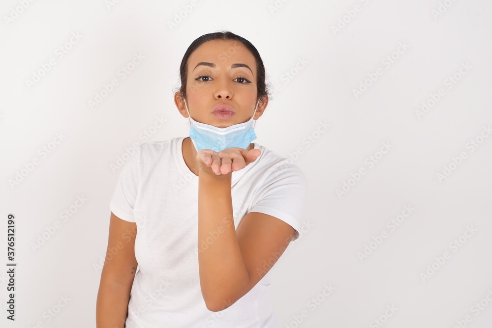 Young arab woman wearing medical mask standing over isolated white background looking at the camera blowing a kiss with hand on air being lovely and sexy. Love expression.