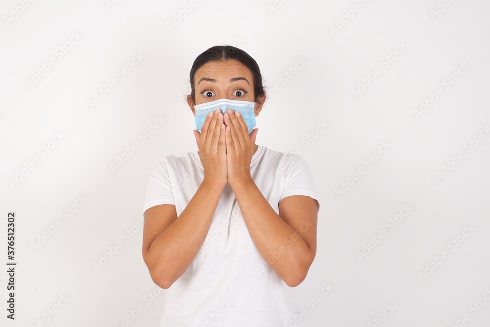 Young arab woman wearing medical mask standing over isolated white background, giggles joyfully, covers mouth, has fun alone, has natural laughter, hears positive story or funny anecdote Young arab woman wearing medical mask standing over isolated white background, giggles joyfully, covers mouth, has fun alone, has natural laughter, hears positive story or funny anecdote