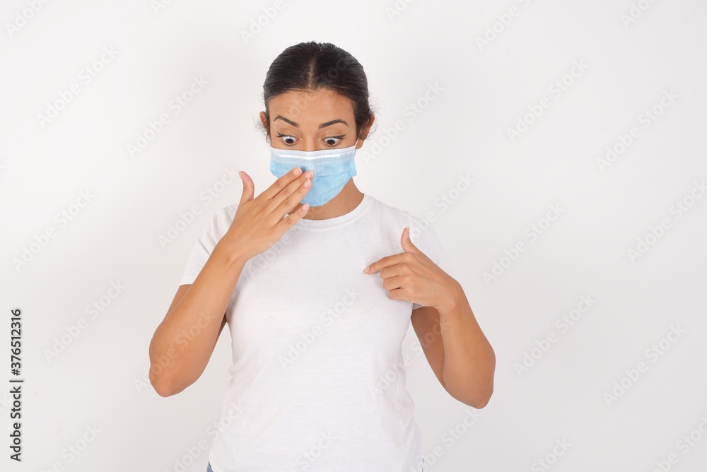 Shocked Young arab woman wearing medical mask standing over isolated white wall look surprisedly down, indicates at blank with fore finger, . Scared female finds out price on usual shirt Shocked Young arab woman wearing medical mask standing over isolated white wall look surprisedly down, indicates at blank with fore finger, . Scared female finds out price on usual shirt