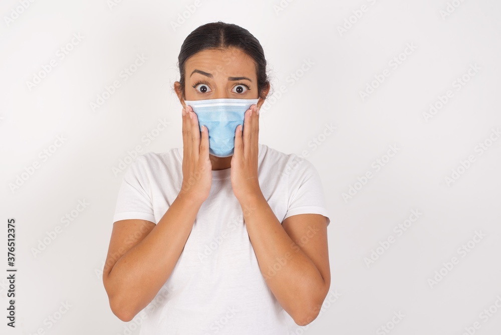 Studio shot of scared terrified Young arab woman wearing medical mask standing over isolated white wall shocked with prices at shop, being short of money to buy something, People and human emotions 