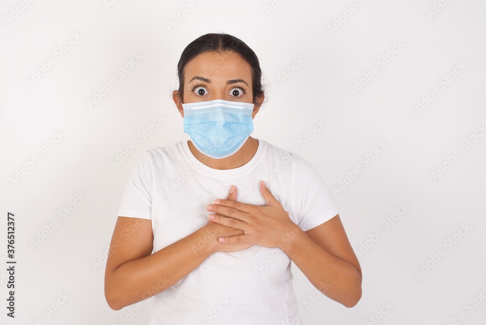 Scared Young arab woman wearing medical mask standing looks with frightened expression, keeps hands on chest, being puzzled to notice something strange, People, hush reaction and emotions. Scared Young arab woman wearing medical mask standing looks with frightened expression, keeps hands on chest, being puzzled to notice something strange, People, hush reaction and emotions.