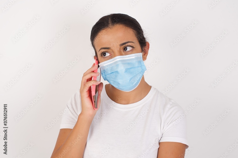 Sad Young arab woman wearing medical mask standing over isolated white background talking on smartphone. Communication concept. Sad Young arab woman wearing medical mask standing over isolated white background talking on smartphone. Communication concept.