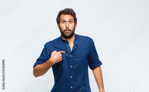 young latin handsome man feeling confused, puzzled and insecure, pointing to self wondering and asking who, me? photo