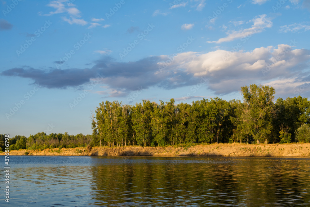 Sunset on the river. Steep river shore, overgrown with poplar grove. The sandy beach is highlighted by the rays of setting sun