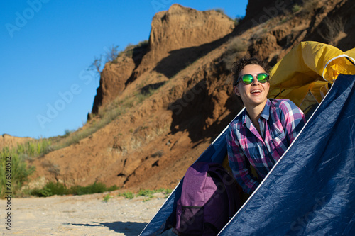 Happy young brunette woman tourist in sunglasses peeping out of touristic tent, smiling looking into distance, enjoying vacation adventure in camping on mountains and blue sky background