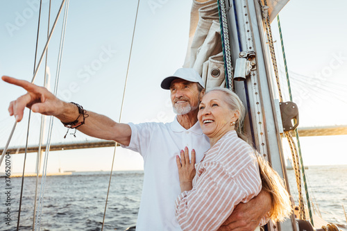 Mature man pointing into the distance and hugging his wife. Two smiling people enjoying a boat trip at sunset.