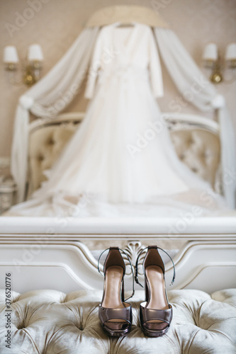 Close up of fashionable shoes on high heel and blurred wedding dress on background. Beautiful bridal clothing elements. Luxury footwear for important formal occasion. Concept of old fashioned style.