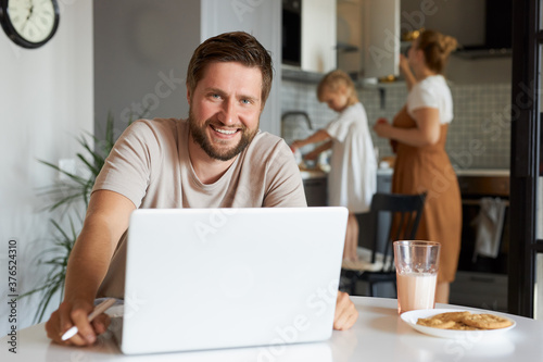 portrait of smiling man working from home, handsome guy look at camera, sit with laptop. family in the background
