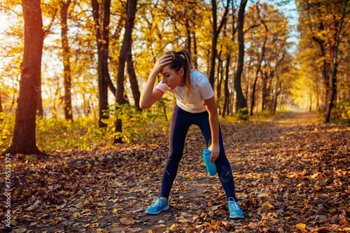 Runner having rest after workout in autumn forest. Tired woman holding water bottle. Sportive active lifestyle