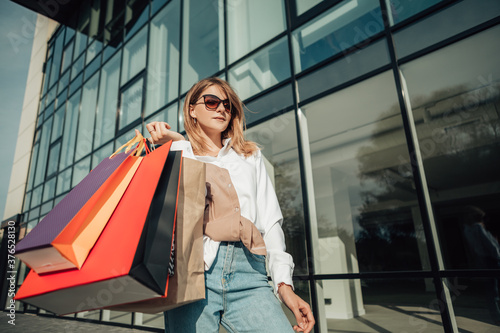 Portrait of european lady hold hand colorful bags woman after shopping in sunglasses white shirt jeans retail store near mall on the street sales black friday season bokeh background
