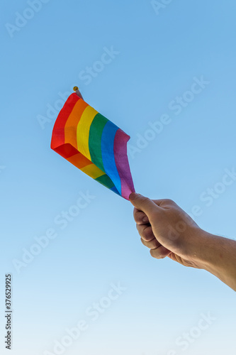 Male hand holding flag of LGBT collective, clear sky background.
