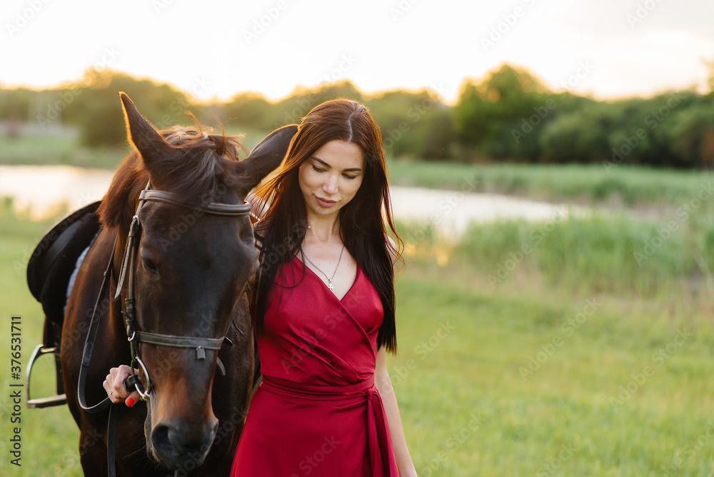 Fototapeta premium A young pretty girl in a red dress poses on a ranch with a thoroughbred stallion at sunset. Love and care for animals