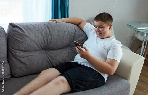 young fat boy chatting with friend, sit with mobile phone, teen boy in domestic wear sits on sofa and look at smartphone, smile. leisure time