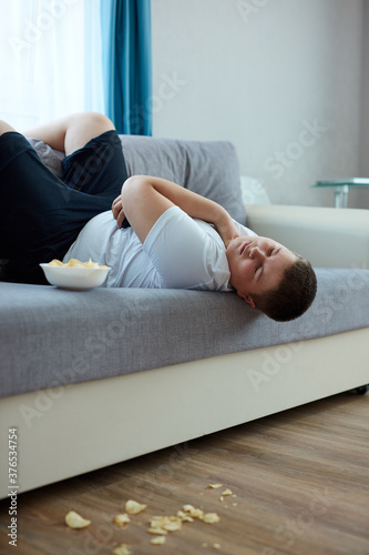 overeat fat boy sleep on sofa in living room, young caucasian teen boy fall asleep while he was watching tv and eating crisps