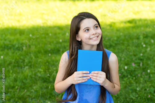 Little happy girl reading book outdoors sunny day, daydreamer concept