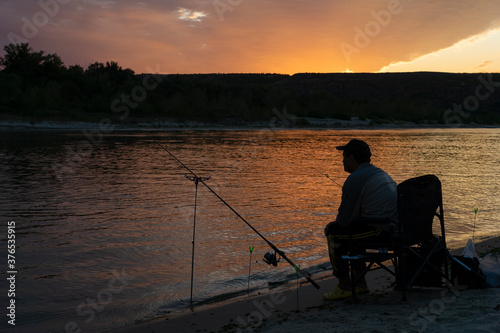 silhouette of fisherman in sunset sitting on bank of river with fishing rod in summer evenong and looking over river, relaxing time in scenery outdoors. Way of calm down concept
