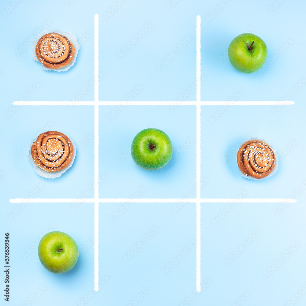 Green apples vs cinnamon buns in tic tac toe or noughts and crosses game, healthy vs unhealthy food concept, top view, square