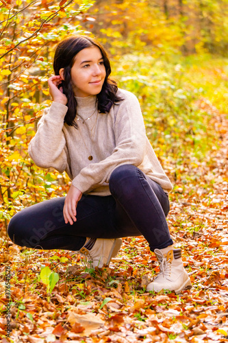 Young pretty stylish dressed girl with beige sweater and black jeans sits in the colorful autumn forest in October and brushes her hair behind the ear