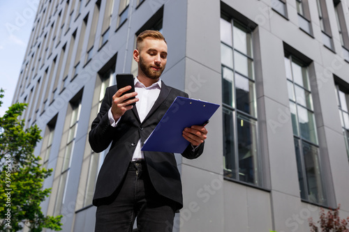 Portrait of smiling young businessman dressed in formal clothes standing by glass building reading report in open folder holding phone