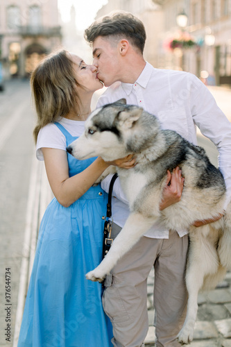 Lovely Caucasian couple in love, dressed in stylish casual wear, hugging and kissing outdoors in the city, having fun and spending time together with their husky dog