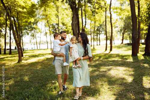 Loving parents with little kids walking at the park, adorable mother hold in arms beautiful daughter, cute son sitting in fathers hands, enjoy happy family weekend concept © shunevich