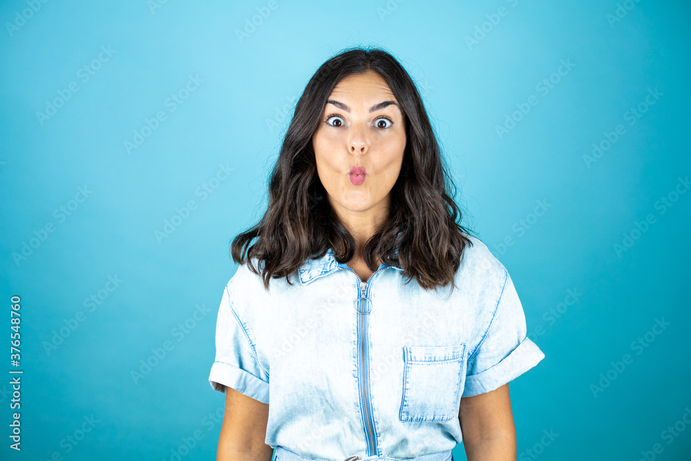 Young beautiful woman wearing a denim jumpsuit over isolated blue background making fish face with lips, crazy and comical gesture Young beautiful woman wearing a denim jumpsuit over isolated blue background making fish face with lips, crazy and comical gesture