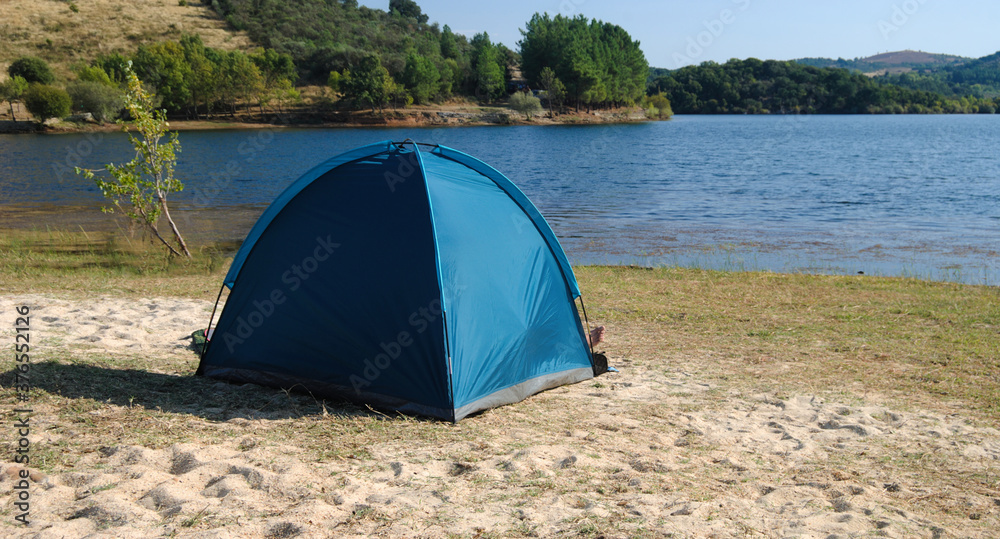 Tenda de campismo azul montada numa praia fluvial ou albufeira numa montanha, campista dentro da tenda com um pé de fora da tenda, Macedo de Cavaleiros