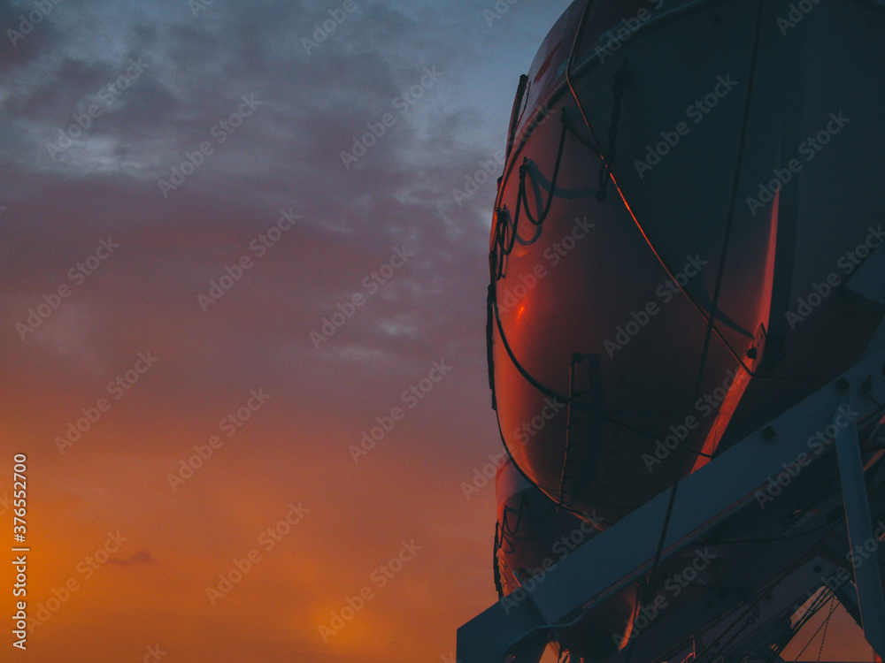Detail of a lifeboat on a cruise ship or ferry at a romantic evening sunset setting. Rescue lifeboats high up on a crane, ready to be deployed. Detail of a lifeboat on a cruise ship or ferry at a romantic evening sunset setting. Rescue lifeboats high up on a crane, ready to be deployed.