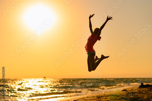 Happy girl jumping on the beach against the background of the sea and sunset. Sunset at sea. Girl jumping raising her hands up.