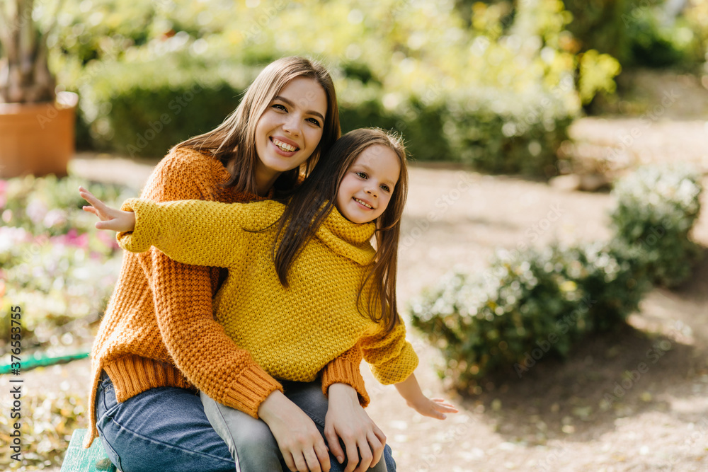Fototapeta premium Inspired little girl playing with mom in park. Cheerful young woman sitting on nature background with daughter.