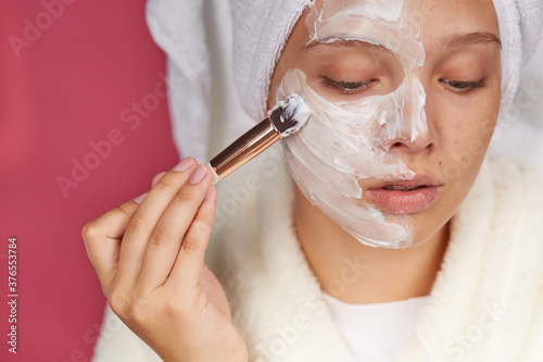 A beautiful young woman applies a face mask to her face with a brush. A young woman in a Terry dressing gown and towel on an isolated red background