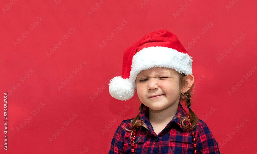 little girl winks one eye in a red Santa hat on a red background. portrait. Merry Christmas and Happy Holidays. little girl winks one eye in a red Santa hat on a red background. portrait. Merry Christmas and Happy Holidays.