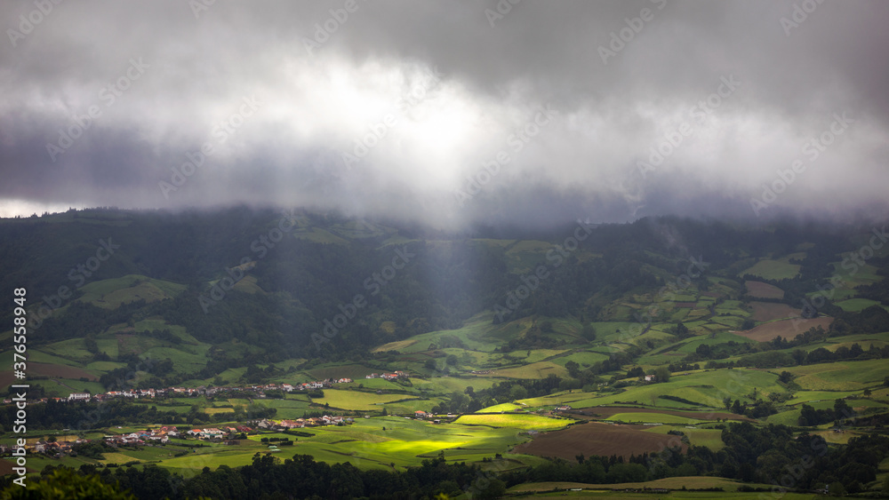 Fototapeta premium Bright sunlight shining through hole of clouds to dark scene of mountain range, Azores, Sao Miguel, Portugal. The beam of light through the dark clouds on the mountains in Azores, Portugal.