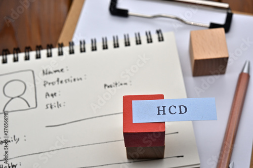 A note with the user's persona written on it is placed on a clipboard along with a pen. A sticky note stamped with "HCD" is placed on a wooden cube.