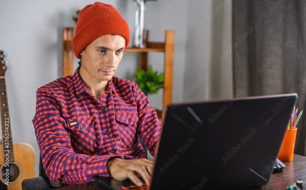Fototapeta premium Modern young man in a red checked shirt and hat is working on a laptop. Concept of distance communication and remote job