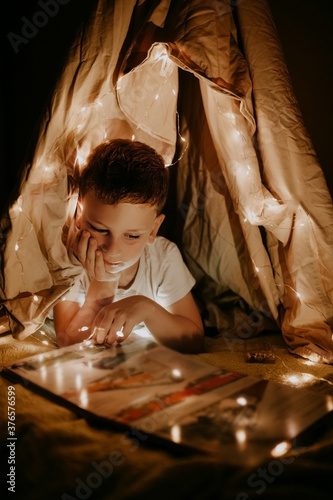 blondy preschooler boy reads a book in hut at home with garlands and lights at night, games for children at home